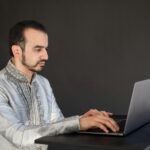 About Middle Eastern man in traditional attire working on a laptop, focused and engaged.